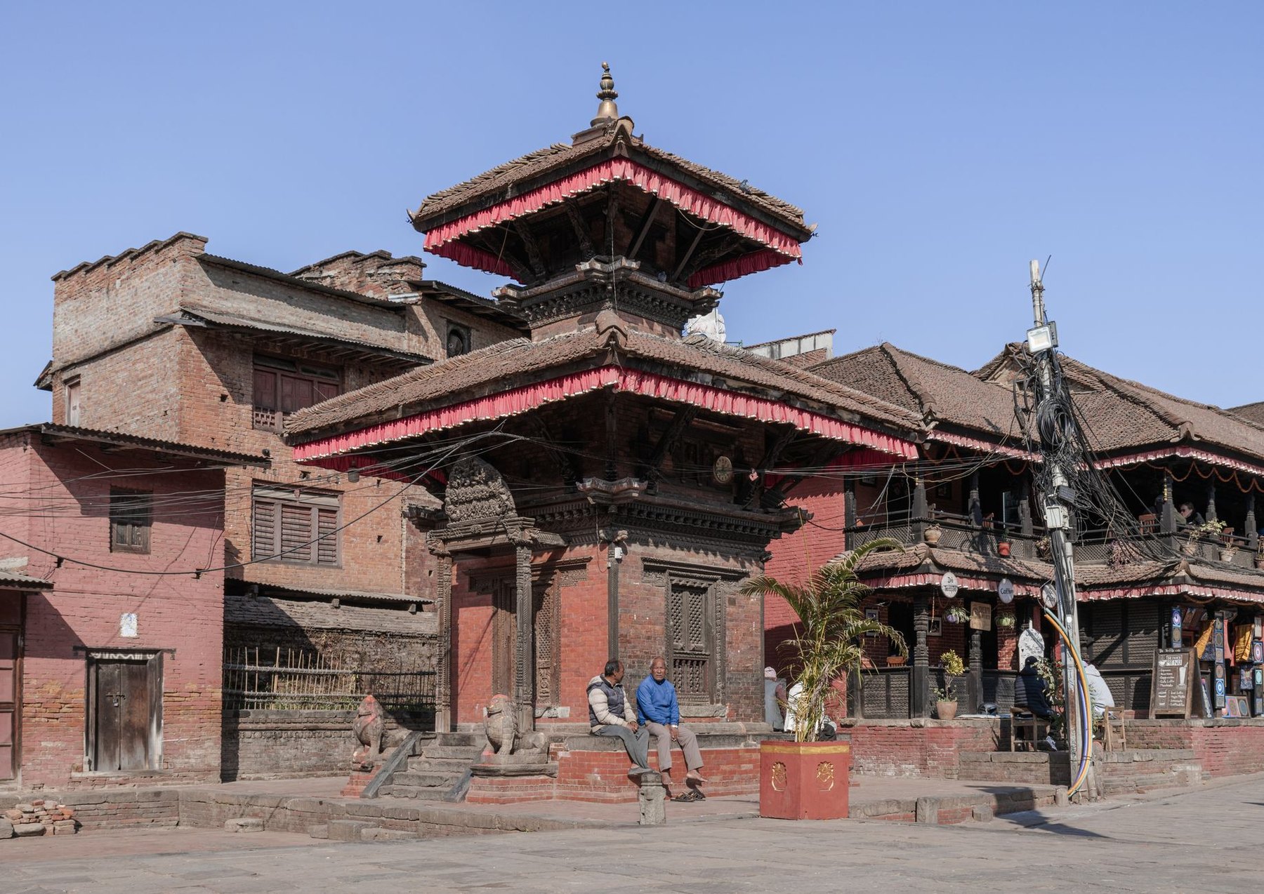Two men sitting on the brick plinth of a tiered Newar temple in Bhaktapur Durbar Square in the morning, with carved wooden struts, shuttered shopfronts and an empty swept square around them.