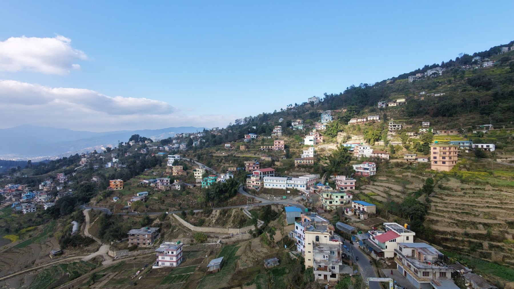 A hillside village on the eastern rim of the Kathmandu Valley, houses and terraces stacked down a green ridge with the hazy valley floor far below.