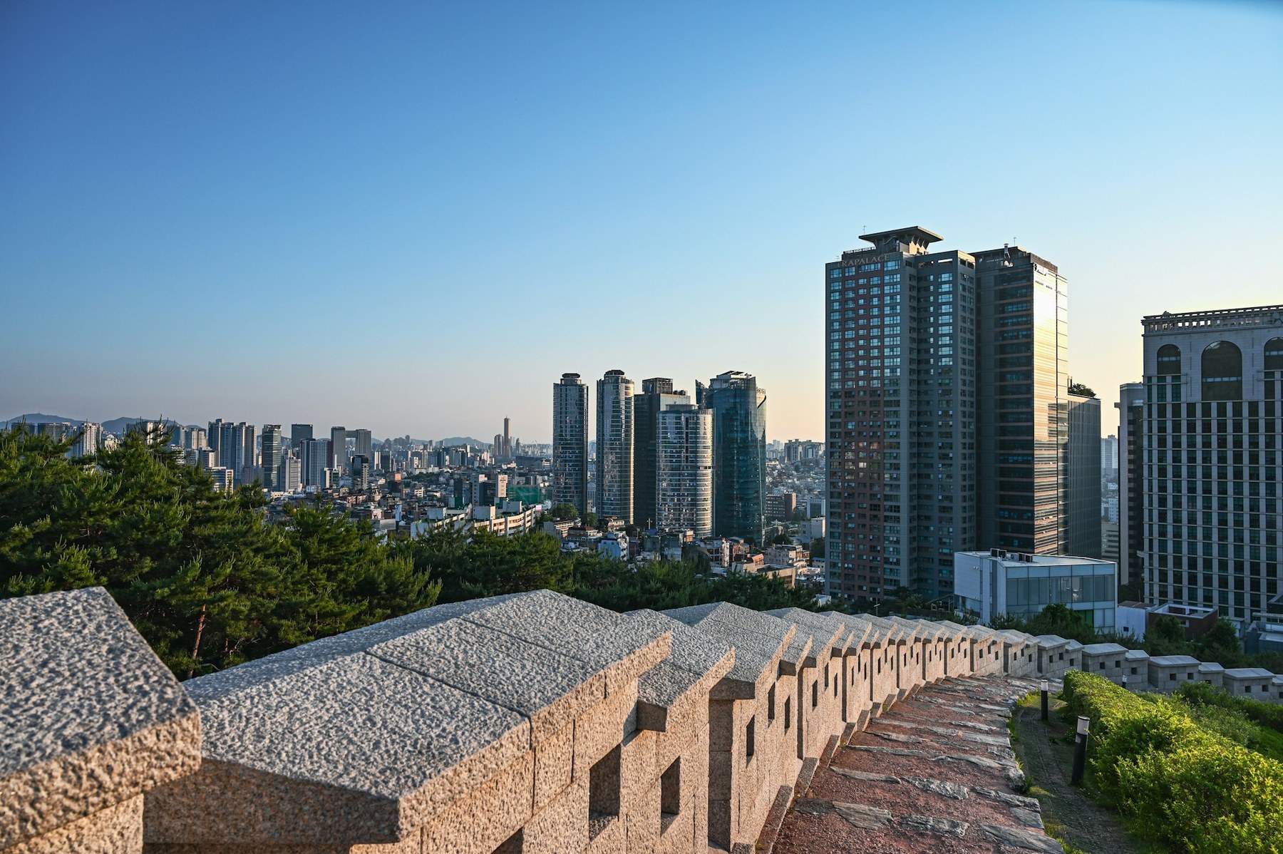 The Seoul City Wall running along a ridge above the city: a crenellated grey granite parapet with pine trees on the left and the high-rise skyline of Seoul beyond.