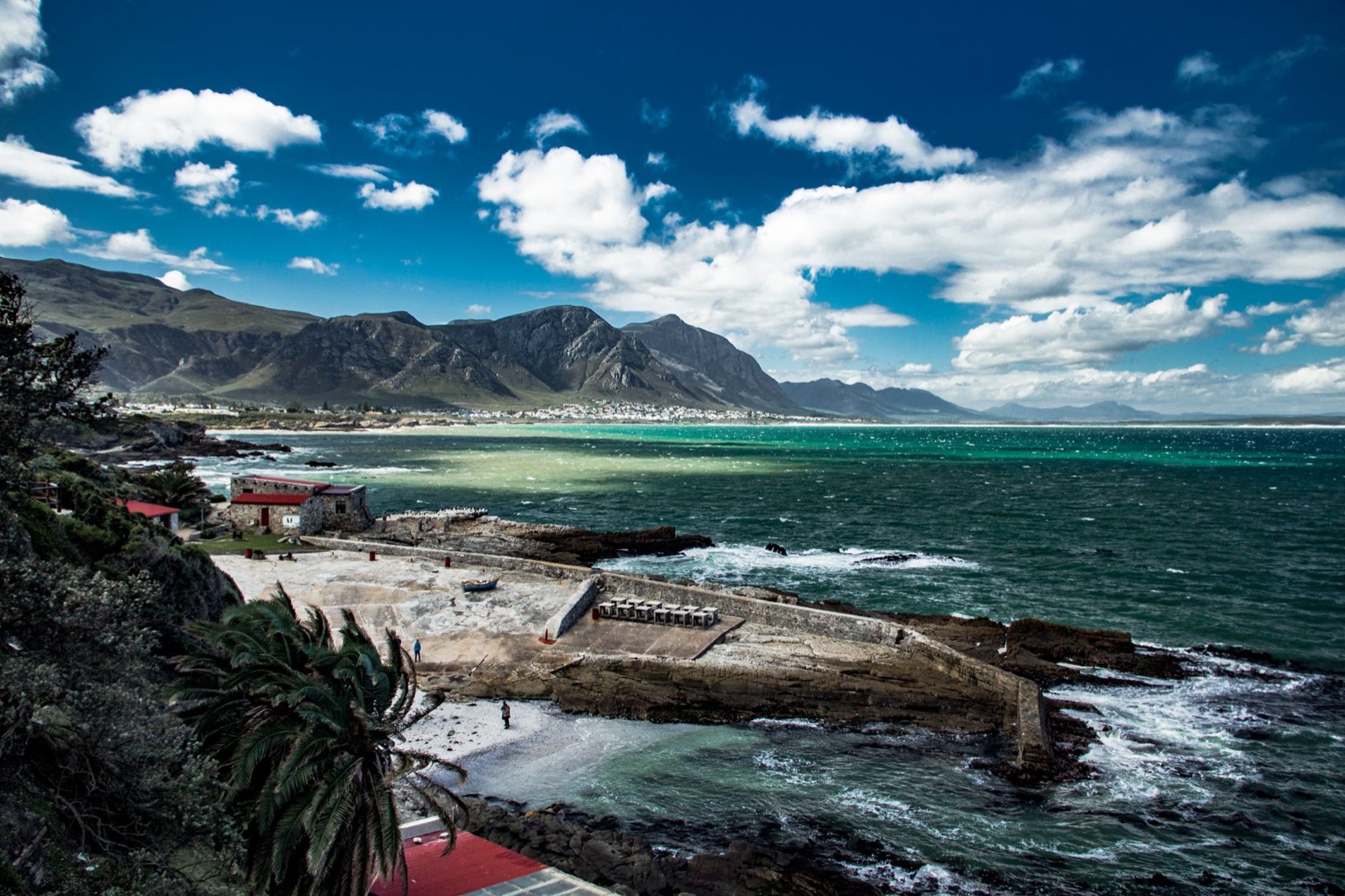 The Old Harbour at Hermanus seen from the cliff path above: a stone slipway and harbour wall below green cliffs, with the turquoise sweep of Walker Bay and the Kleinrivier mountains beyond.